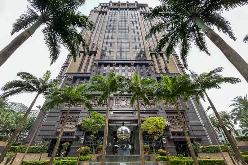 Best Areas to Stay in Singapore 3 Tall modern building with large glass windows, palm trees at the entrance, and symmetrical landscaping in the foreground—one of the top areas to stay for comfort and style.