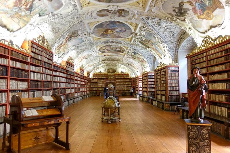 Ornate library hall with arched, frescoed ceilings, tall bookshelves filled with books, wooden floor, and globes and statues placed throughout the space.