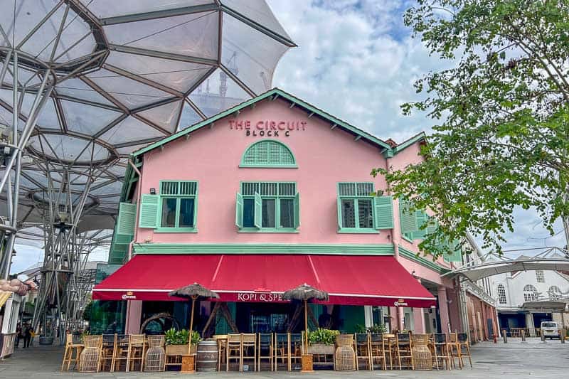 Best Areas to Stay in Singapore 7 A pink two-story building with green shutters, a red awning labeled "Kopi & Spice," and outdoor seating, this spot sits under a large canopy—perfect for exploring popular areas to stay nearby.