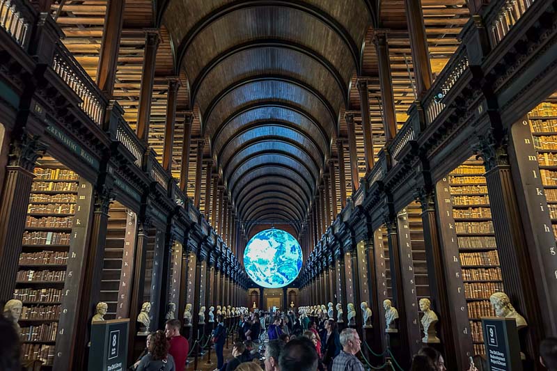 Large library hall with tall bookshelves, arched wooden ceiling, marble busts lining both sides, and a large illuminated globe at the far end. People are walking through the hall.