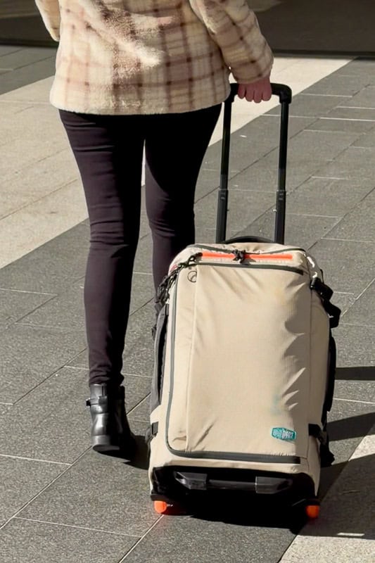BAGSMART BLAST Series Review: Tested on My Travels 13 A person wearing a checked jacket and black trousers pulls a beige rolling suitcase with orange wheels along a pavement, while carrying the versatile Blast 40L Rucksack for extra storage.