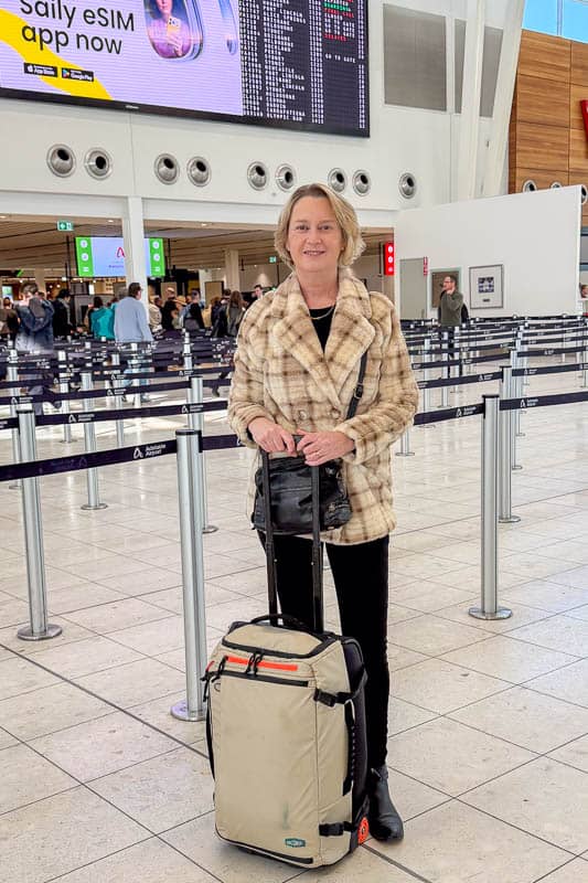 BAGSMART BLAST Series Review: Tested on My Travels 14 A woman wearing a checked coat stands in an airport terminal, holding the handle of a beige suitcase and carrying a sleek Blast 40L Rucksack. There are black queue barriers, people in the background, and a large digital flight information board overhead.