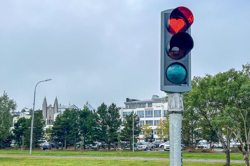 10 Days in Iceland: Our Real Itinerary 37 A traffic light shows a red heart-shaped signal against a cloudy sky, with trees, parked cars, and buildings in the background—capturing the charm you might spot during 10 days in Iceland.