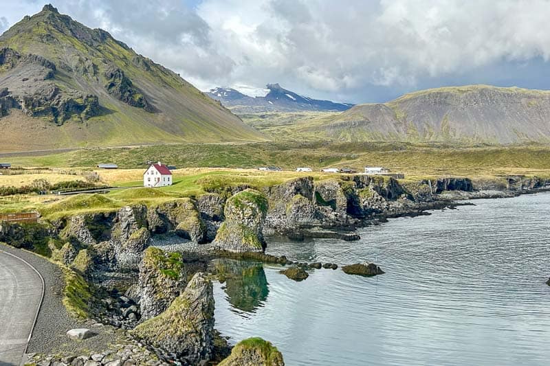 10 Days in Iceland: Our Real Itinerary 46 A white house sits on grassy cliffs overlooking calm water, with rugged mountains and scattered clouds in the background—a tranquil scene you might encounter during 10 days in Iceland.