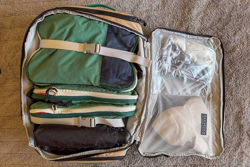 BAGSMART BLAST Series Review: Tested on My Travels 4 An open suitcase on a carpeted floor shows neatly packed green and beige packing cubes, a rolled black item—possibly the Blast 40L Rucksack—and a mesh compartment containing a light-coloured hat and some folded clothes.