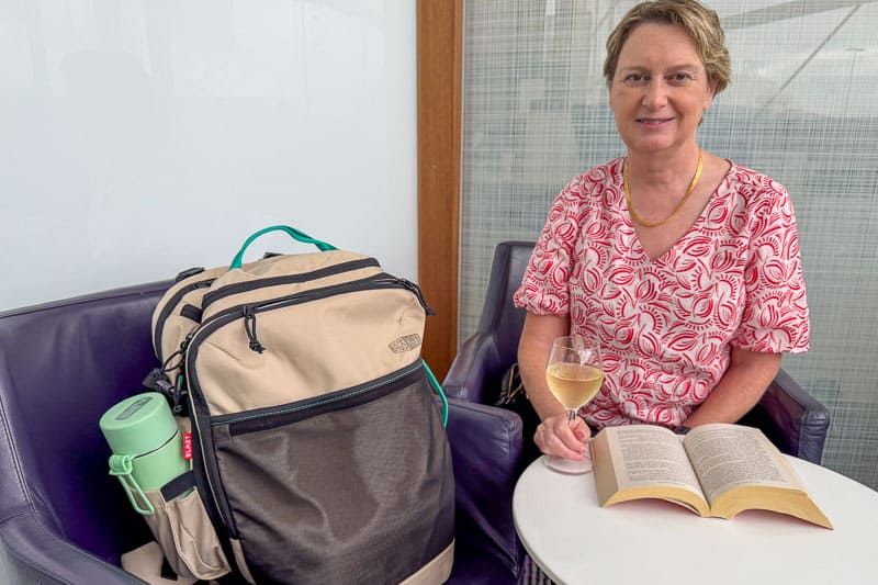 BAGSMART BLAST Series Review: Tested on My Travels 10 A woman sits in a chair holding a glass of white wine. Next to her is a beige and grey Blast 40L Rucksack with a green water bottle in the side pocket. An open book lies on the small round table in front of her.