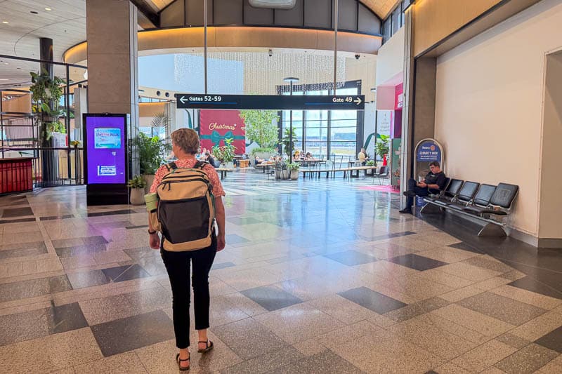 BAGSMART BLAST Series Review: Tested on My Travels 9 A traveller carrying a Blast 40L Rucksack walks through an airport terminal towards gate signs, with seating to the right and a Chick-fil-A restaurant and plants visible in the distance.