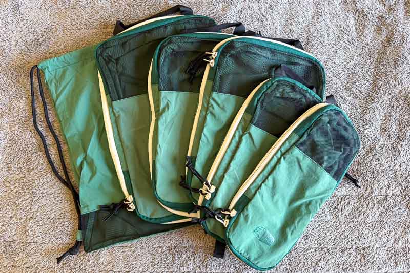 BAGSMART BLAST Series Review: Tested on My Travels 8 Four green packing cubes and a green drawstring bag are arranged on a textured beige surface. The packing cubes vary in size, with black mesh panels and light-coloured zips.