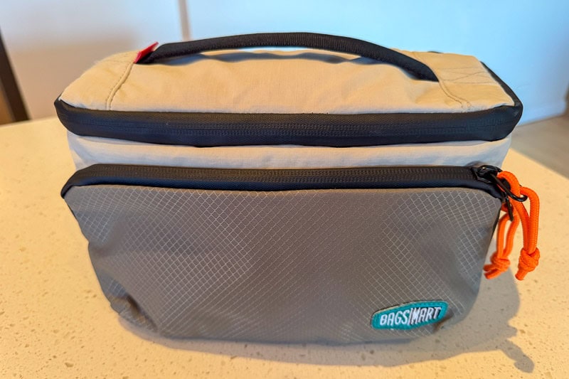 BAGSMART BLAST Series Review: Tested on My Travels 15 A grey and beige BAGSMART zippered pouch with a handle on top, an orange zip pull, and a front pocket—perfect for organising essentials inside your Blast 40L Rucksack—sits on a speckled worktop.
