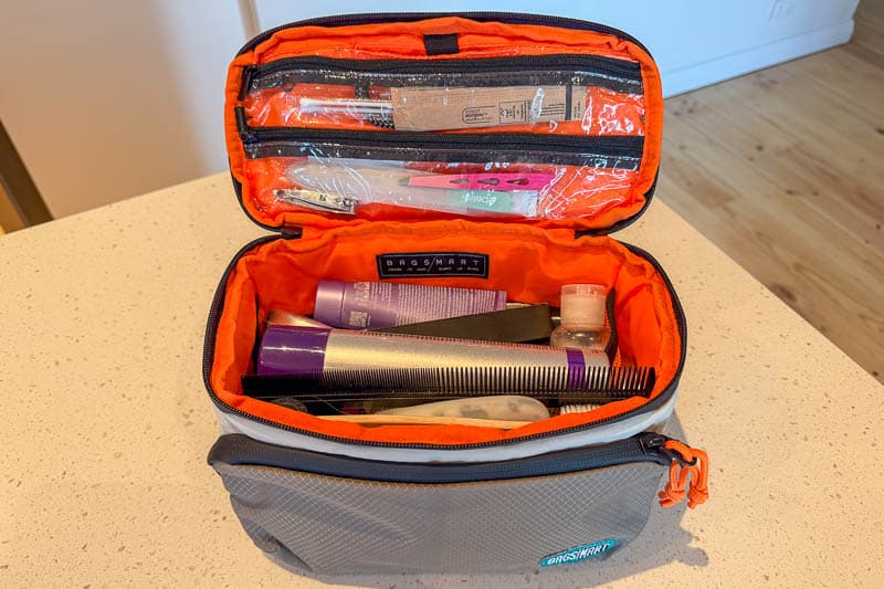 BAGSMART BLAST Series Review: Tested on My Travels 16 An open grey and orange toiletry bag sits on a countertop next to the Blast 40L Rucksack. The bag contains items such as a comb, toothpaste, a toothbrush, a razor, bottles, and various small toiletries in the main and top compartments.