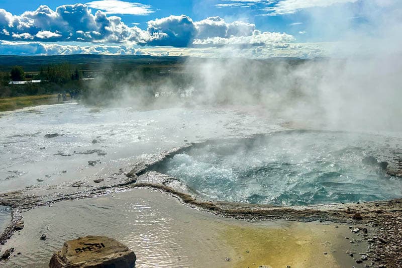 Things to Know Before Going to Iceland 5 A steaming geothermal hot spring with bubbling water and mineral deposits, basking under a partly cloudy Icelandic sky. In the foreground, a stone labelled "BLESI" welcomes those going to Iceland to witness this natural wonder.