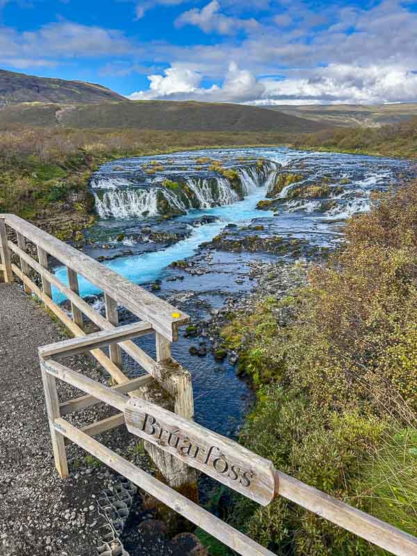 5 Must-See Waterfalls in Iceland 5 A wooden railing with a sign reading “Bruarfoss” overlooks a blue waterfall flowing over rocky terrain, surrounded by autumn foliage under a partly cloudy sky.