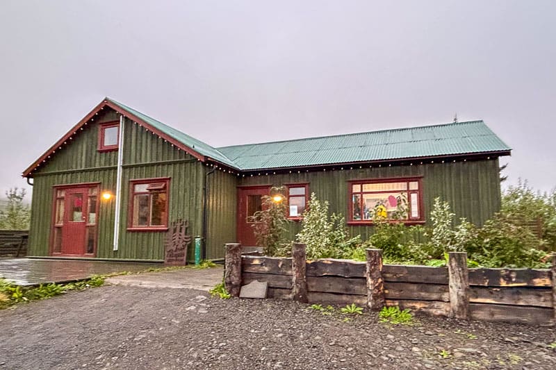 Iceland Campgrounds 12 A green wooden house with a metal roof, front porch lights on, and plants along the front, photographed on an overcast day.