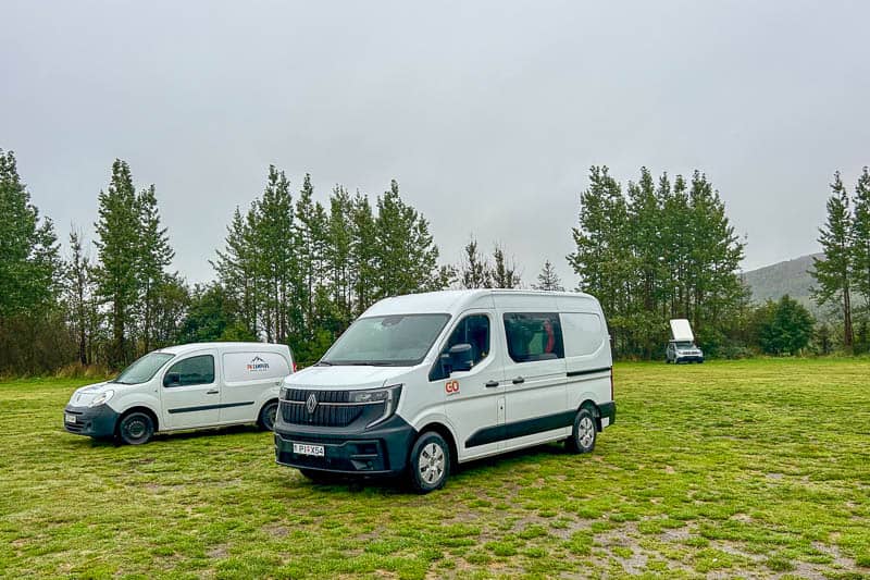 Iceland Campgrounds 13 Two white vans are parked on a grassy field with trees in the background under an overcast sky. A third vehicle is visible with its roof tent open in the distance.