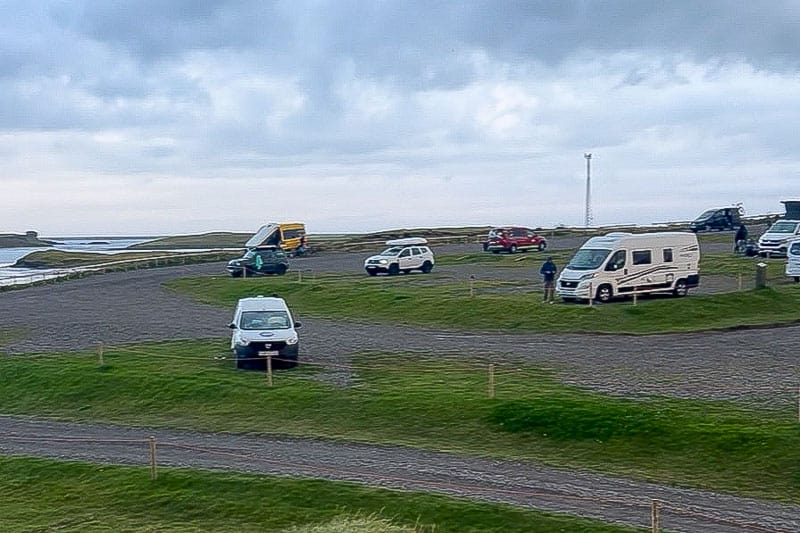 Iceland Campgrounds 9 Several vehicles, including cars and camper vans, are parked on a grassy, tiered Iceland Campgrounds site near the coastline under a cloudy sky.
