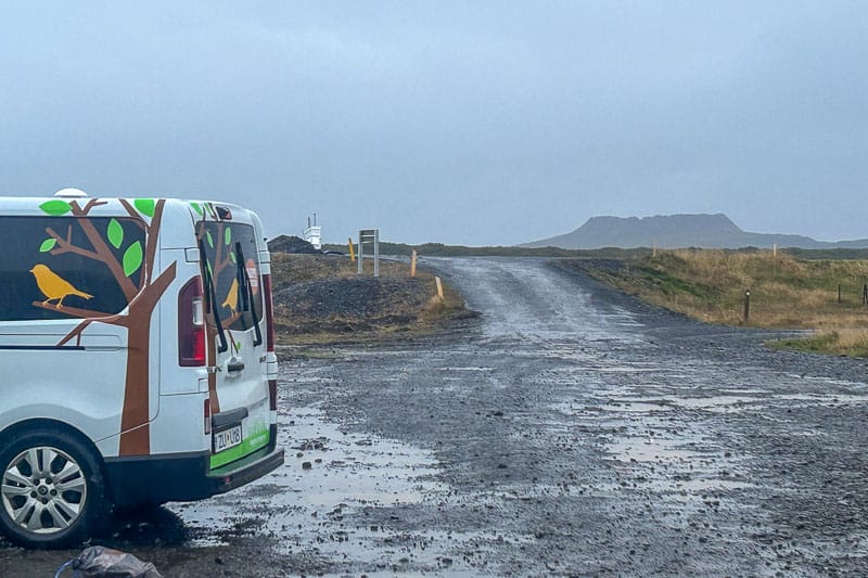 Iceland Campgrounds 15 A van with bird and tree graphics is parked on a wet, gravel road with grassy fields and a distant mountain under an overcast sky.
