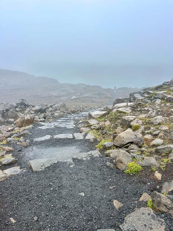 5 Must-See Waterfalls in Iceland 22 A rocky path with uneven stones leads downhill through a foggy, barren landscape. Large rocks and sparse patches of green vegetation are scattered along the sides of the track. Visibility is low due to thick fog.