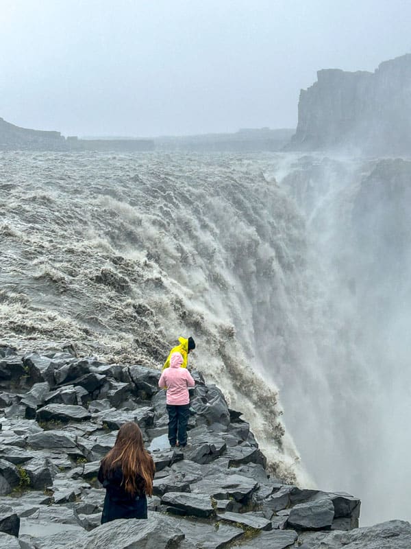 5 Must-See Waterfalls in Iceland 24 Two people, one adult and one child in raincoats, stand on rocky ground near the edge of a large, misty waterfall under a grey, overcast sky.