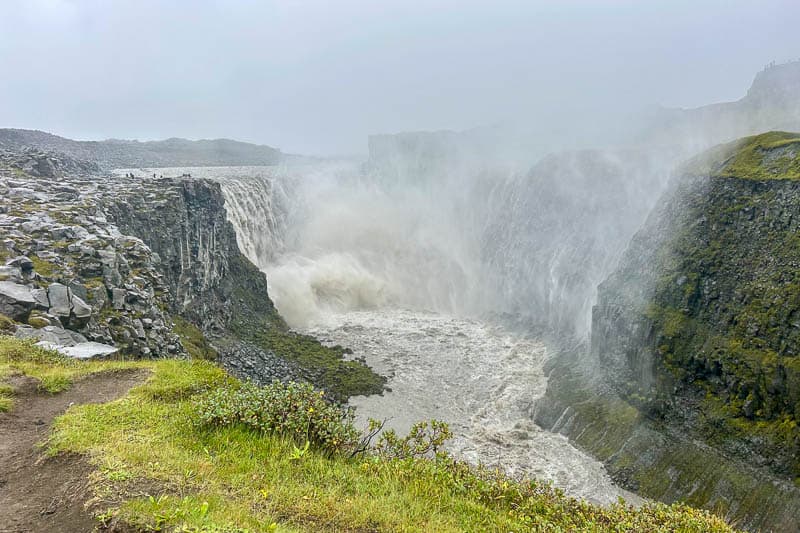 5 Must-See Waterfalls in Iceland 21 A large waterfall cascades into a deep rocky gorge with mist rising, surrounded by green vegetation and overcast skies—a breathtaking scene you might witness during 10 days in Iceland.