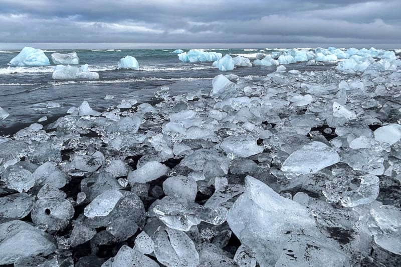 Things to Know Before Going to Iceland 3 Large chunks of ice scattered along a black sand beach with more ice floating in the sea under a cloudy sky—an unforgettable sight when going to Iceland.