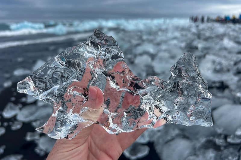 10 Days in Iceland: Our Real Itinerary 25 A hand holds a jagged, clear chunk of ice with more ice pieces scattered on a rocky shore, capturing just one magical moment from 10 days in Iceland as people explore in the distant background.