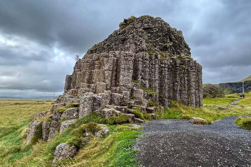 10 Days in Iceland: Our Real Itinerary 20 Large basalt rock formation with hexagonal columns rises from grassy landscape under a cloudy sky; a gravel path curves nearby—a must-see for anyone spending 10 days in Iceland.