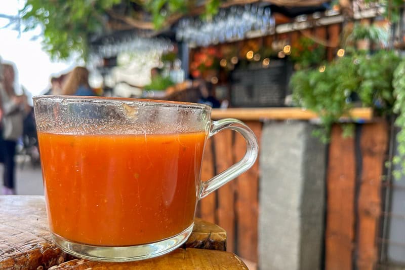 Driving the Golden Circle in Iceland 7 A clear Golden Circle glass cup filled with tomato soup sits on a wooden surface. The background shows an out-of-focus bar area with hanging glasses and greenery, creating a casual, rustic atmosphere.