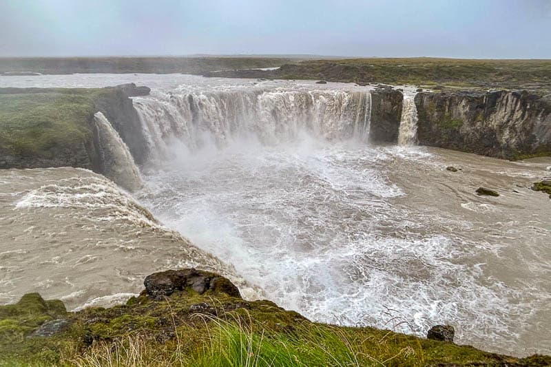 The Diamond Circle: Iceland's Other Scenic Drive 12 A wide, powerful waterfall cascades over a rocky cliff into a turbulent river, surrounded by mossy green terrain under a cloudy sky—an unforgettable sight during 10 days in Iceland.