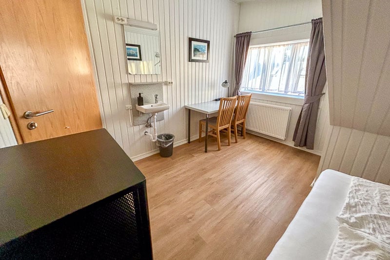 Guesthouse Aurora Review - Our One-Night Stay in Reykjavik 5 A small, tidy room with wooden flooring, a single bed, a wall-mounted washbasin, a mirror, a desk with two wooden chairs, a window with curtains, and a radiator under the window.