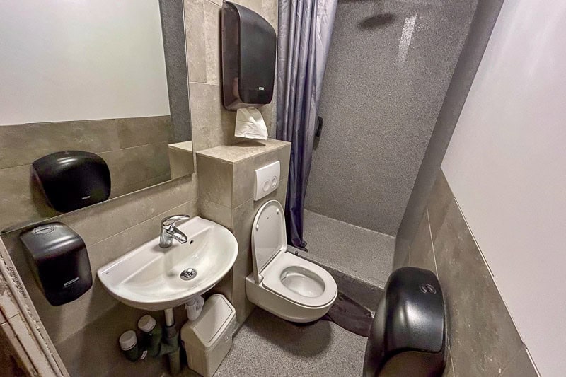 Guesthouse Aurora Review - Our One-Night Stay in Reykjavik 6 A small bathroom with a wall-mounted basin, an open toilet, soap and paper towel dispensers, a mirror, and a shower area separated by a curtain. The floor and walls are covered in grey tiles.
