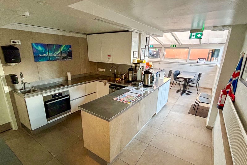 Guesthouse Aurora Review - Our One-Night Stay in Reykjavik 7 A modern kitchen with beige tiled floors, white cupboards, stainless steel appliances, a coffee station, a small dining area with three chairs, and a flag hanging on the wall. A window lets in natural light.