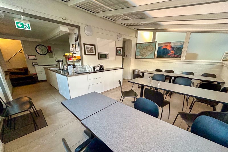 Guesthouse Aurora Review - Our One-Night Stay in Reykjavik 8 A small canteen with several black chairs and grey tables, a counter with coffee and tea supplies, a wall clock, and framed pictures on white walls under a bright ceiling light.