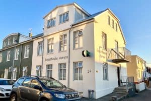 Guesthouse Aurora Review - Our One-Night Stay in Reykjavik 46 A three-storey cream-coloured building labelled “Guesthouse Aurora,” with a side staircase and parked cars along the street, is pictured on a clear day. The neighbouring building is green.
