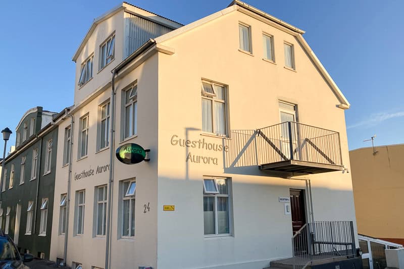 Guesthouse Aurora Review - Our One-Night Stay in Reykjavik 3 A beige two-storey building with the words "Guesthouse Aurora" on the wall, featuring multiple windows, a balcony, and a small sign reading "Aurora" next to the entrance. The building is lit by sunlight.