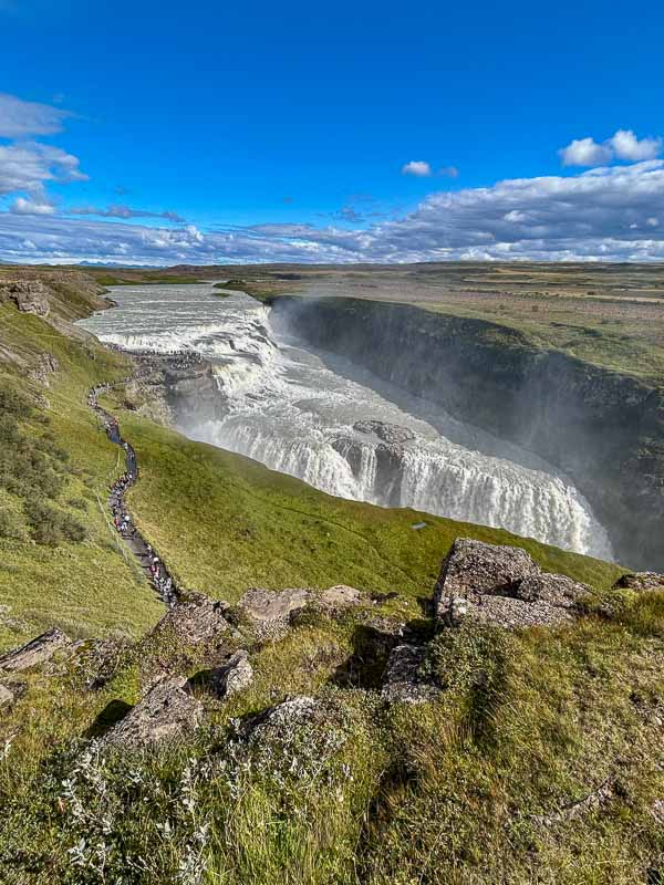 5 Must-See Waterfalls in Iceland 7 A wide waterfall cascades down rocky cliffs surrounded by green grassy hills under a bright blue sky with scattered clouds. A path with people walking follows the edge of the cliffs beside the waterfall.