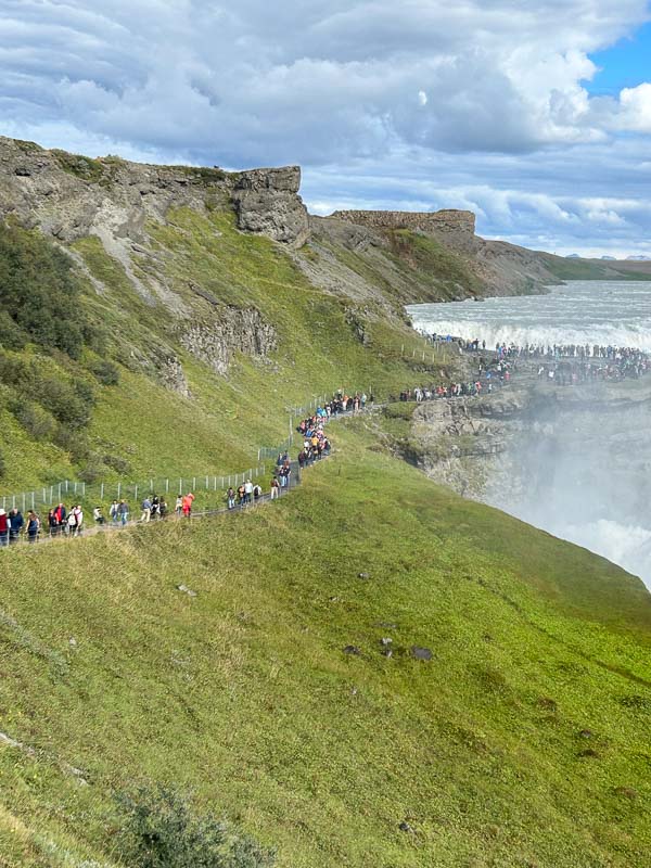5 Must-See Waterfalls in Iceland 8 A crowd of people walks along a fenced path on a grassy hillside next to a waterfall. Rocky cliffs rise in the background under a partly cloudy sky. Mist from the waterfall drifts over the lower right side of the image.