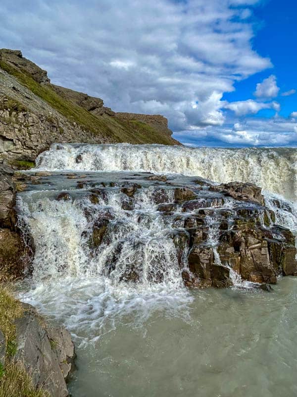 5 Must-See Waterfalls in Iceland 9 A small waterfall flows over layered rocks, surrounded by grassy slopes and rocky cliffs under a partly cloudy blue sky. White water splashes into a shallow, pale pool below.