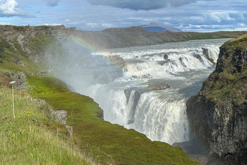 5 Must-See Waterfalls in Iceland 6 A wide waterfall cascades through a rocky gorge with mist creating a faint rainbow; grassy cliffs and a group of people—perhaps on a 10 days in Iceland adventure—are visible nearby under a partly cloudy sky.