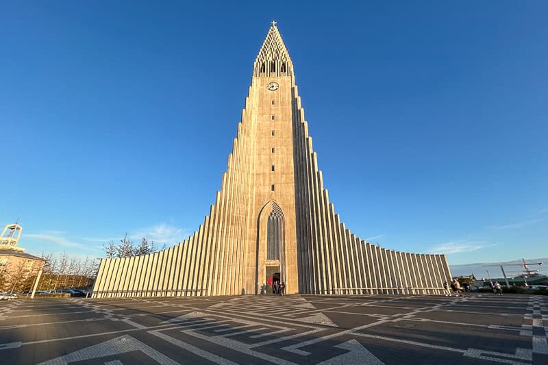 European Churches Worth Visiting 12 A tall, concrete church with a stepped design and a central clock tower stands under a clear blue sky—an iconic sight you won't want to miss during 10 days in Iceland.