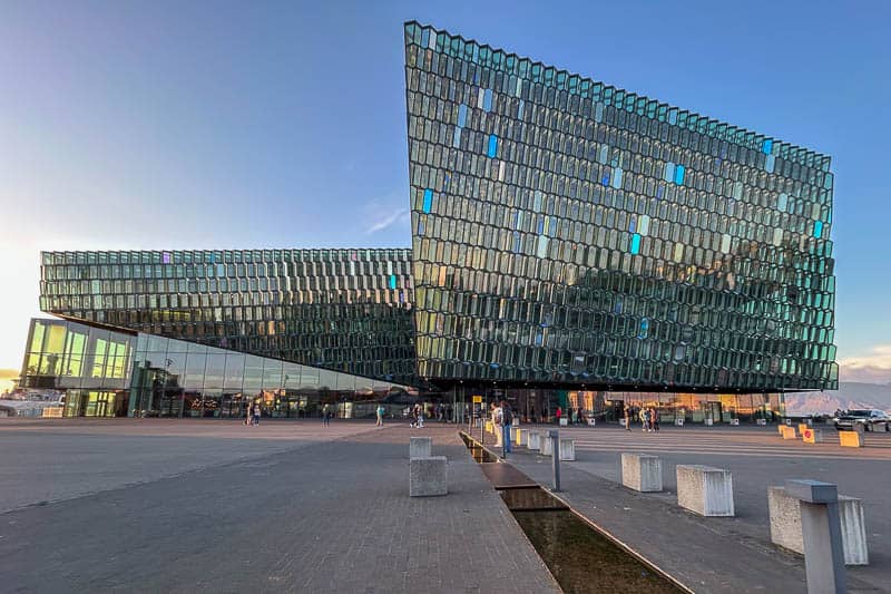 Things to Know Before Going to Iceland 4 A modern glass building with an angular, geometric design stands under a clear sky. Several people, perhaps dreaming of going to Iceland, walk near its entrance. The façade reflects sunlight and sky, creating a shimmering effect.