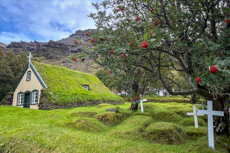10 Days in Iceland: Our Real Itinerary 23 A small church with a grass-covered roof sits beside a graveyard with white crosses and a tree bearing red berries, set against a hilly landscape—an idyllic scene you might find during 10 days in Iceland.