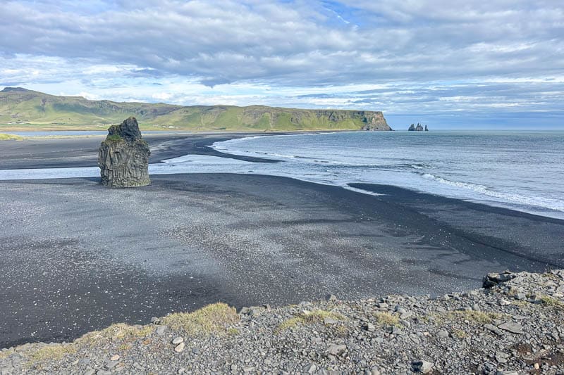 Things to Know Before Going to Iceland 9 A black sand beach with a large rock formation near the shore, gentle waves, and distant cliffs under a partly cloudy sky—perfect scenery for anyone going to Iceland. Grassy hills and mountains rise in the background.