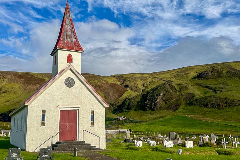 Things to Know Before Going to Iceland 8 A small white church with a red roof and steeple stands near a graveyard, nestled among green grass and hills under a partly cloudy sky—just the kind of scene you might see when going to Iceland.