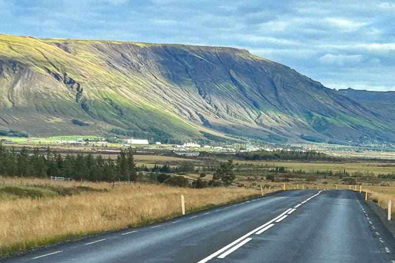 Driving the Golden Circle in Iceland 10 A tarmac road stretches into the distance along the Golden Circle, passing through grassy fields, small trees, and a large rocky mountain in the background under a cloudy sky.