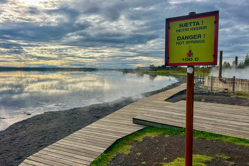 Driving the Golden Circle in Iceland 2 A wooden boardwalk runs along a lake on the Golden Circle, with steam rising in the background. A yellow sign warns in Icelandic and English of hot springs with water at 100°C. The sky is cloudy, with sun reflecting on the water.