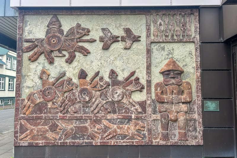 Street Art in Iceland 12 A textured wall mural depicts abstract horsemen with circular shapes, two birds in flight, and a figure sitting with hands on its knees, next to vertical patterns. The art is set in a rectangular frame on an exterior building wall.