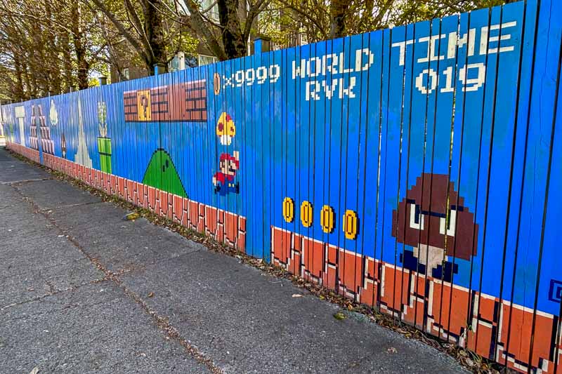 Street Art in Iceland 13 A wooden fence painted with pixel art from Super Mario Bros., featuring Mario, coins, a Goomba, and game stats on a blue background, next to a pavement lined with trees.