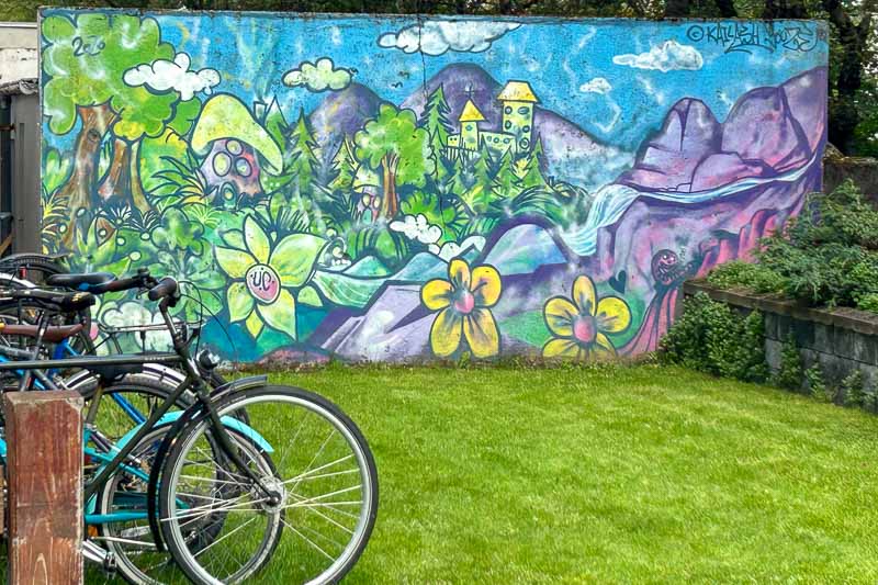 Street Art in Iceland 6 A colourful mural on a wall depicts a whimsical landscape with hills, houses, flowers, and greenery. Several bicycles are parked on the grass in front of the mural.