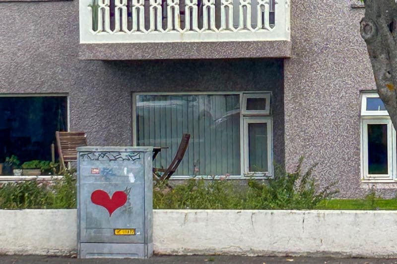Street Art in Iceland 27 A grey utility box with a red heart graffiti stands in front of a residential building with large windows, a balcony, and outdoor chairs behind a low white wall.