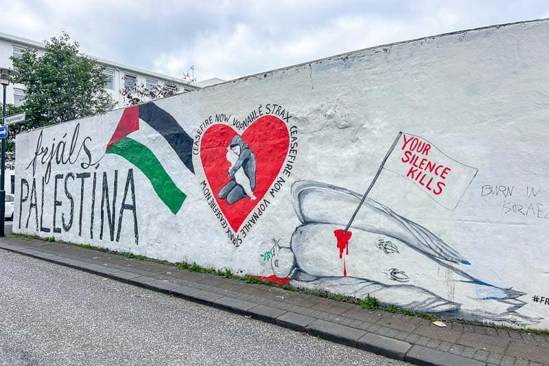 Street Art in Iceland 7 A mural on a white wall shows a dove with an olive branch pierced by a sword labelled "Your silence kills," a red heart with a kneeling figure, and text saying "Free Palestina" and "Ceasefire now." A Palestinian flag is depicted.